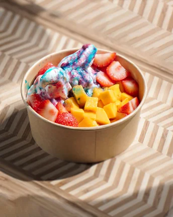 A paper bowl of frozen yogurt topped with mango cubes and sliced strawberries.